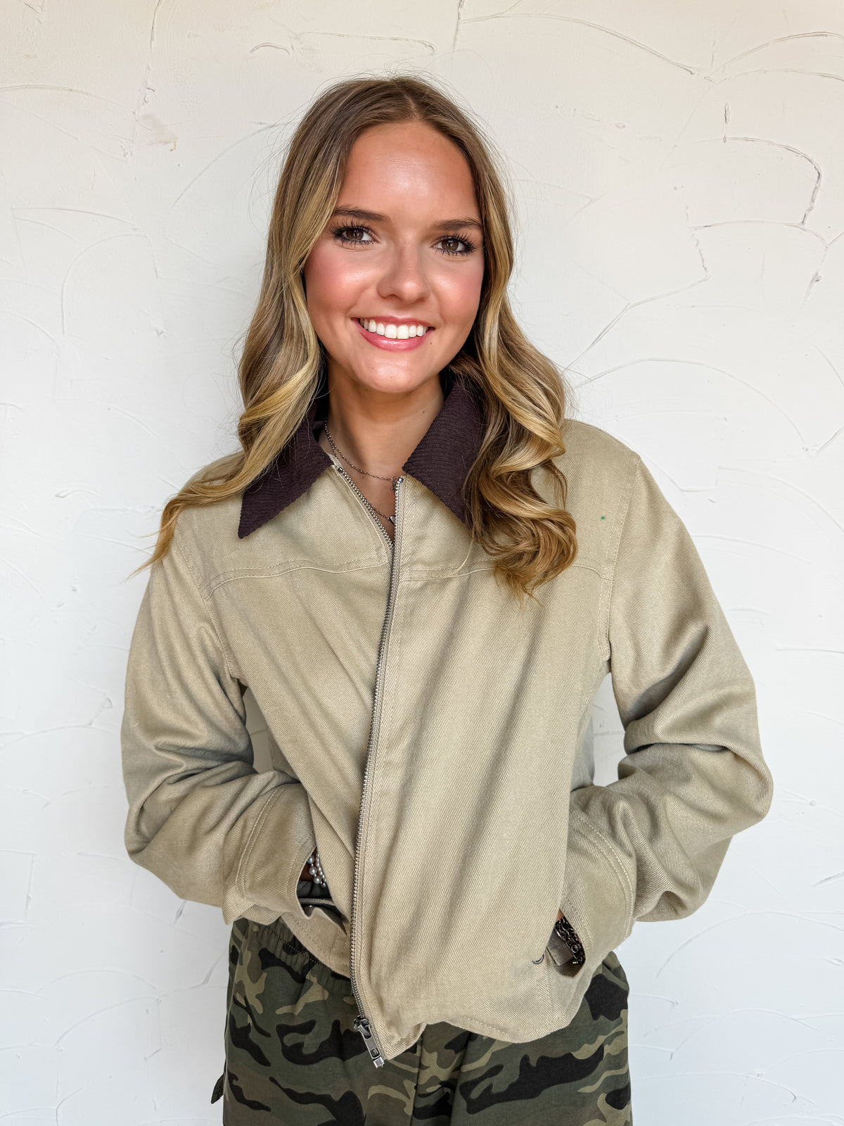 A waist-up photograph of a smiling young woman with long wavy brown hair with blonde highlights posing with her hands in her pockets against a textured white wall. She is wearing a khaki tan canvas full-zip jacket made from 100% cotton, featuring a dark brown corduroy pointed collar and side welt pockets, paired with camouflage print pants, and styled with a delicate silver chain necklace and bracelets.