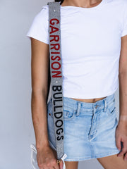 A close-up of a model wearing a white t-shirt and denim skirt, carrying a gray shoulder strap with 'GARRISON BULLDOGS' printed vertically in red and black.