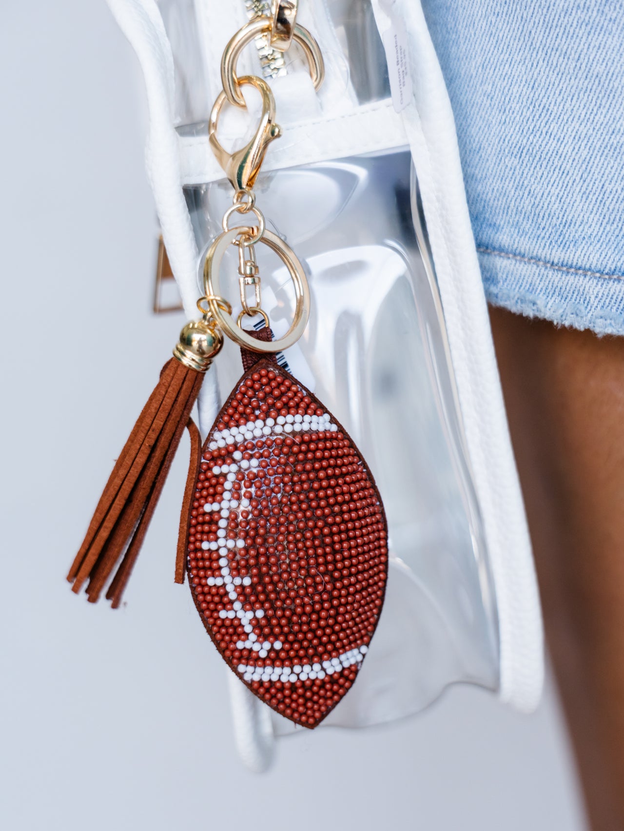 A football-shaped keychain charm with brown leather fringe tassels and gold-tone hardware, featuring a red beaded football body with white beaded laces, shown hanging from a clear handbag.