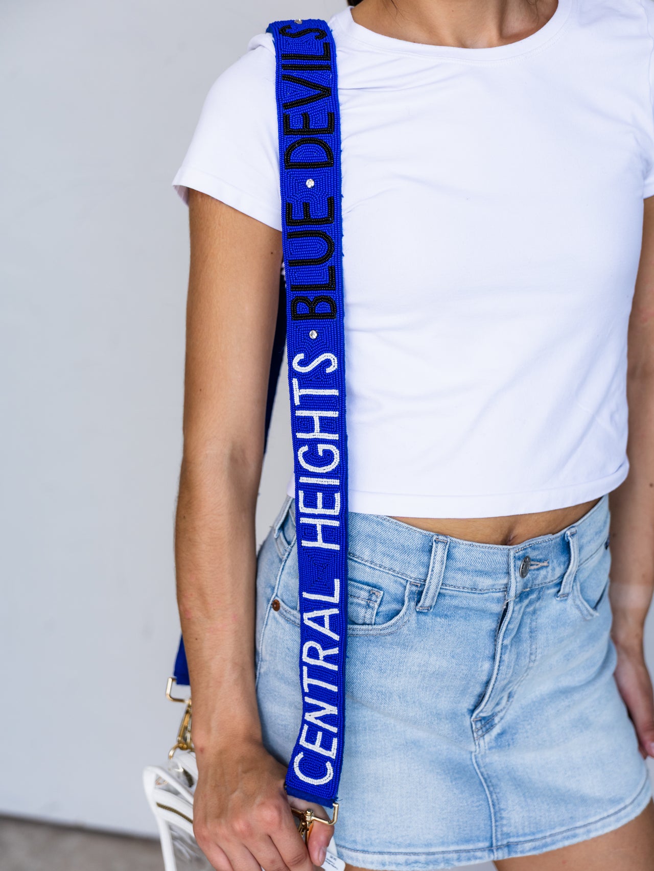 A close-up of a person wearing a white crop top and light blue denim skirt, carrying a white bag with a prominent royal blue shoulder strap printed with the text 'CENTRAL HEIGHTS • BLUE DEVILS'.