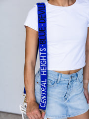 A close-up of a person wearing a white crop top and light blue denim skirt, carrying a white bag with a prominent royal blue shoulder strap printed with the text 'CENTRAL HEIGHTS • BLUE DEVILS'.