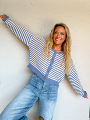 A three-quarter length photograph of a smiling blonde woman with long curly hair posing playfully with arms stretched wide against a textured off-white wall. She is wearing a long-sleeved open-knit cardigan sweater made from a 60% cotton and 40% acrylic blend, featuring horizontal textured stripes in slate blue and cream, a button-down front with brown tortoiseshell buttons, and ribbed blue trim along the neckline, hem, and cuffs, paired with light-wash denim jeans featuring front patch pockets, styled with