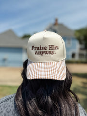 A person viewed from behind wearing a beige trucker-style cap with brown embroidered text reading 'Praise Him anyway.' and a brown-and-white gingham brim, set against a blurred outdoor background of a house and blue sky.