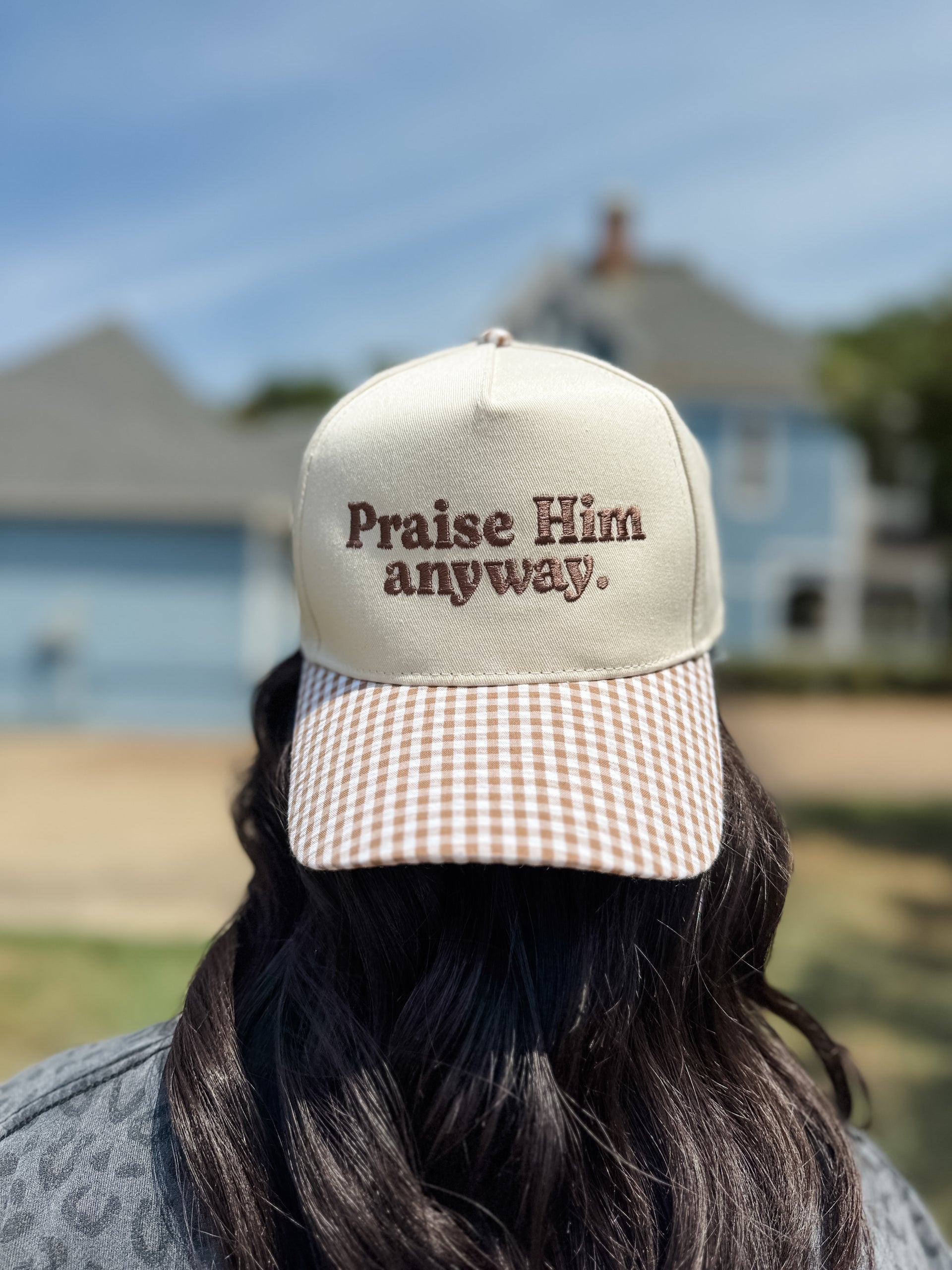 A person viewed from behind wearing a beige trucker-style cap with brown embroidered text reading 'Praise Him anyway.' and a brown-and-white gingham brim, set against a blurred outdoor background of a house and blue sky.