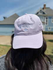 A person viewed from behind wearing a light lavender baseball cap with 'SFASU' embroidered on the front, set against a blurred outdoor background of houses and blue sky.