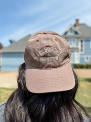 A person viewed from behind wearing a dusty taupe baseball cap with 'NACOGOOCHES 1945' embroidered on the front, set against a blurred background of a blue house and sky.