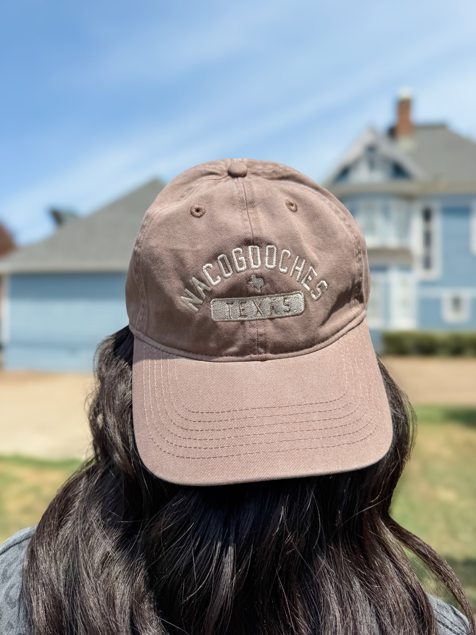 A person viewed from behind wearing a dusty taupe baseball cap with 'NACOGOOCHES 1945' embroidered on the front, set against a blurred background of a blue house and sky.