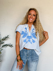 A three-quarter length photograph of a smiling blonde woman with long curly hair holding a section of her hair while posing against a textured white wall next to an olive branch. She is wearing a short-sleeved top made from a 55% cotton and 45% polyester blend, featuring vertical blue and white stripes with bright blue floral embroidery across the chest and blue scalloped trim along the split V-neckline, front-tucked into light-wash denim jeans with patch pockets, styled with a brown leather belt, a gold ch