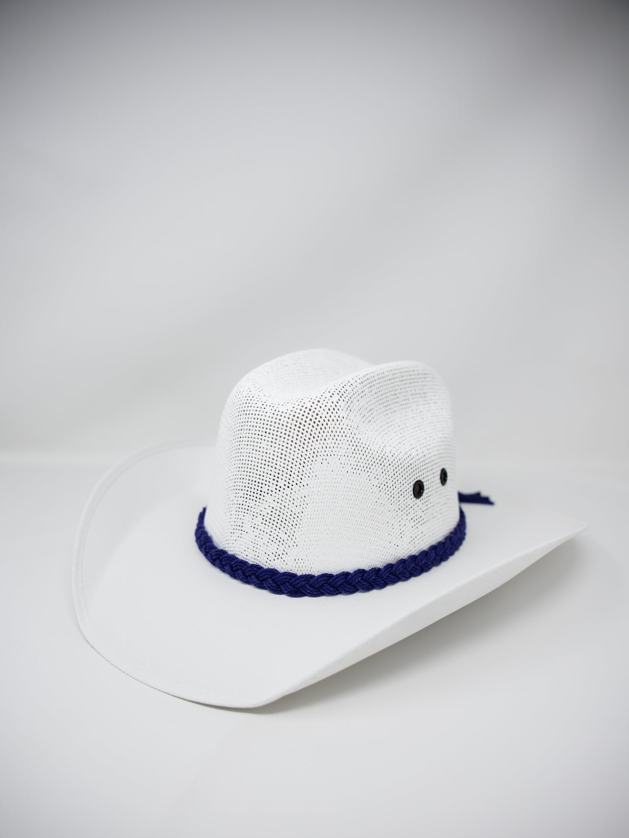 A white woven cowboy hat with a dark blue braided band and small black embroidered eyes, photographed on a light gray studio background.