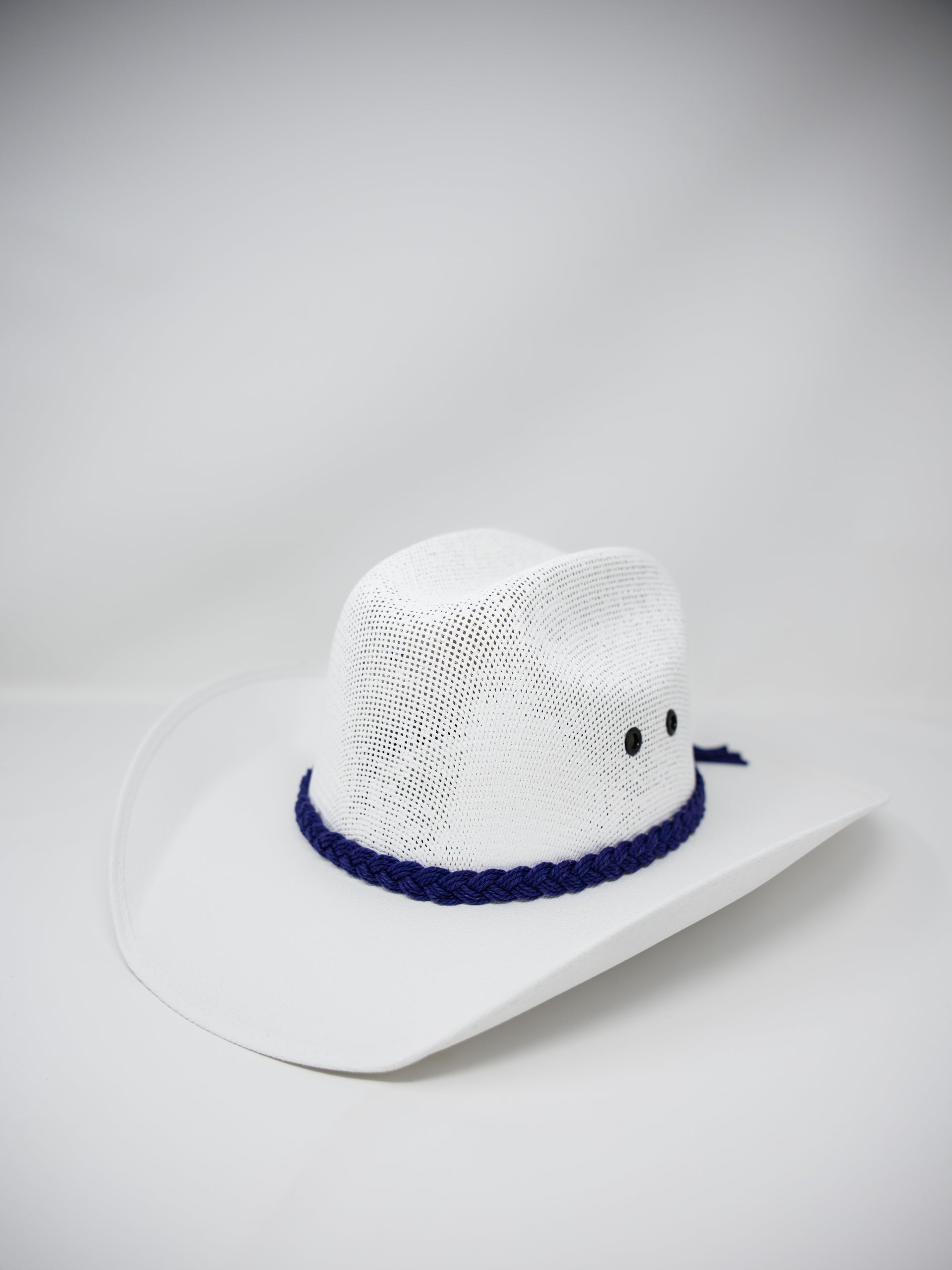 A white woven cowboy hat with a dark blue braided band and small black embroidered eyes, photographed on a light gray studio background.