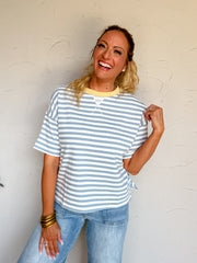 A three-quarter length photograph of a laughing blonde woman with curly ponytail looking toward the camera while holding her shoulder and posing against a textured white wall. She is wearing an oversized short-sleeved t-shirt made from a 60% cotton and 40% polyester blend, featuring horizontal light blue and white stripes, a contrasting pastel yellow ribbed crew neckline, a white stitched triangle V-notch detail, and dropped shoulders, paired with light-wash blue jeans with front patch pockets, styled with 