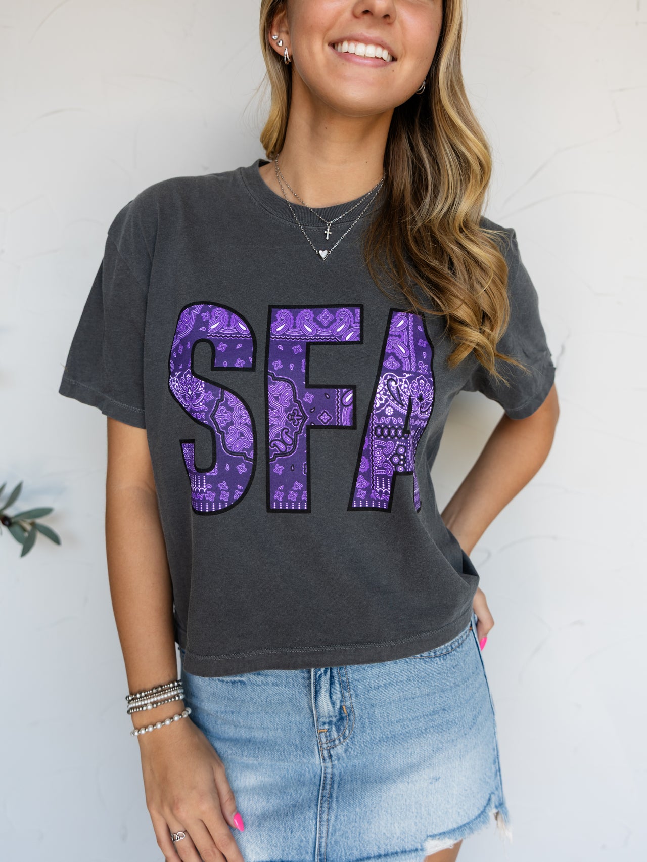 A woman wearing a dark charcoal gray t-shirt with the letters 'SFA' printed in purple paisley pattern, paired with a denim skirt. 100% Cotton