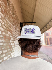 Person wearing a white cap with purple text, standing against a brick wall.