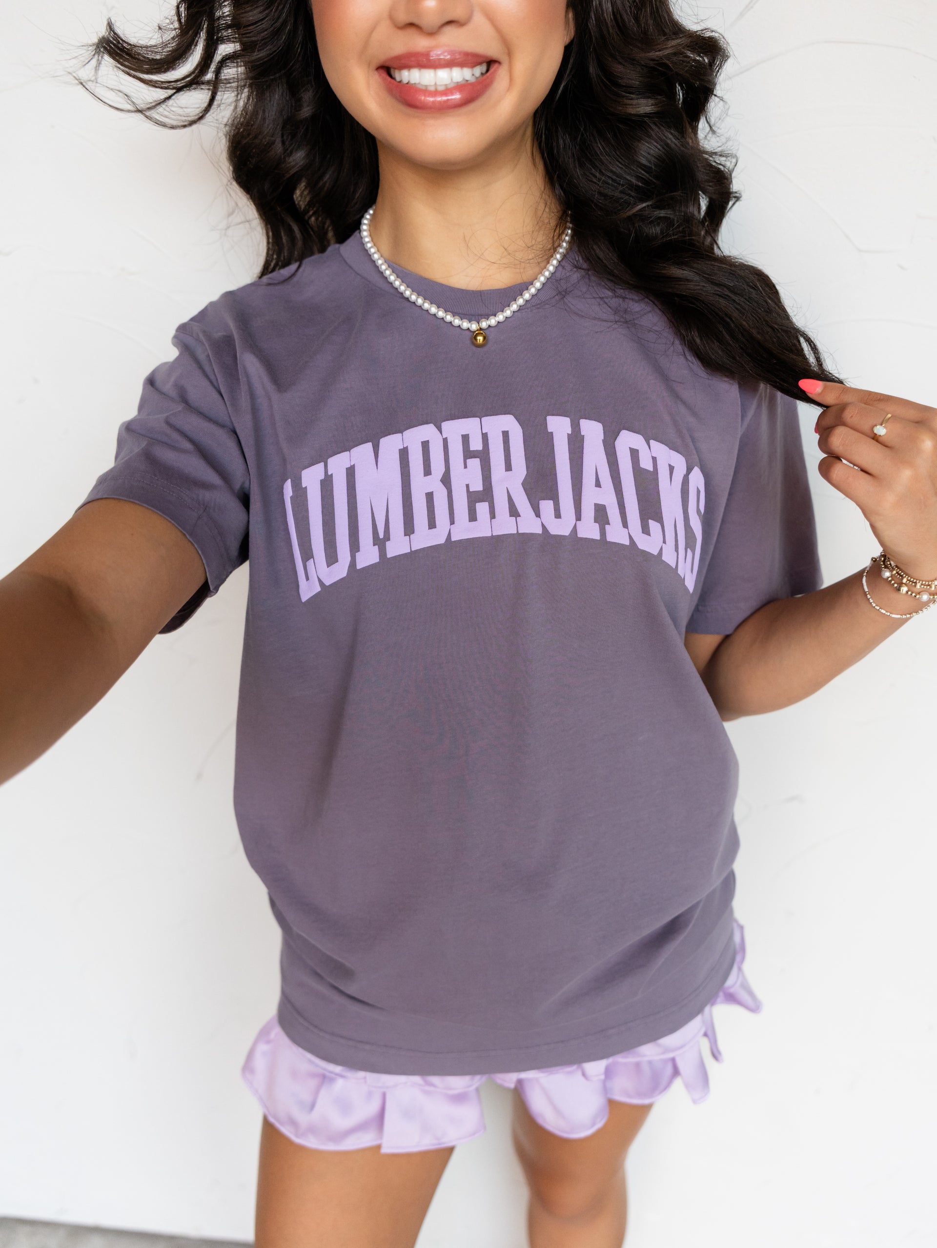 A woman wearing a lavender-purple short-sleeve t-shirt with 'LUMBERJACKS' printed in arched light purple letters across the chest, paired with matching ruffled shorts, against a plain white wall.