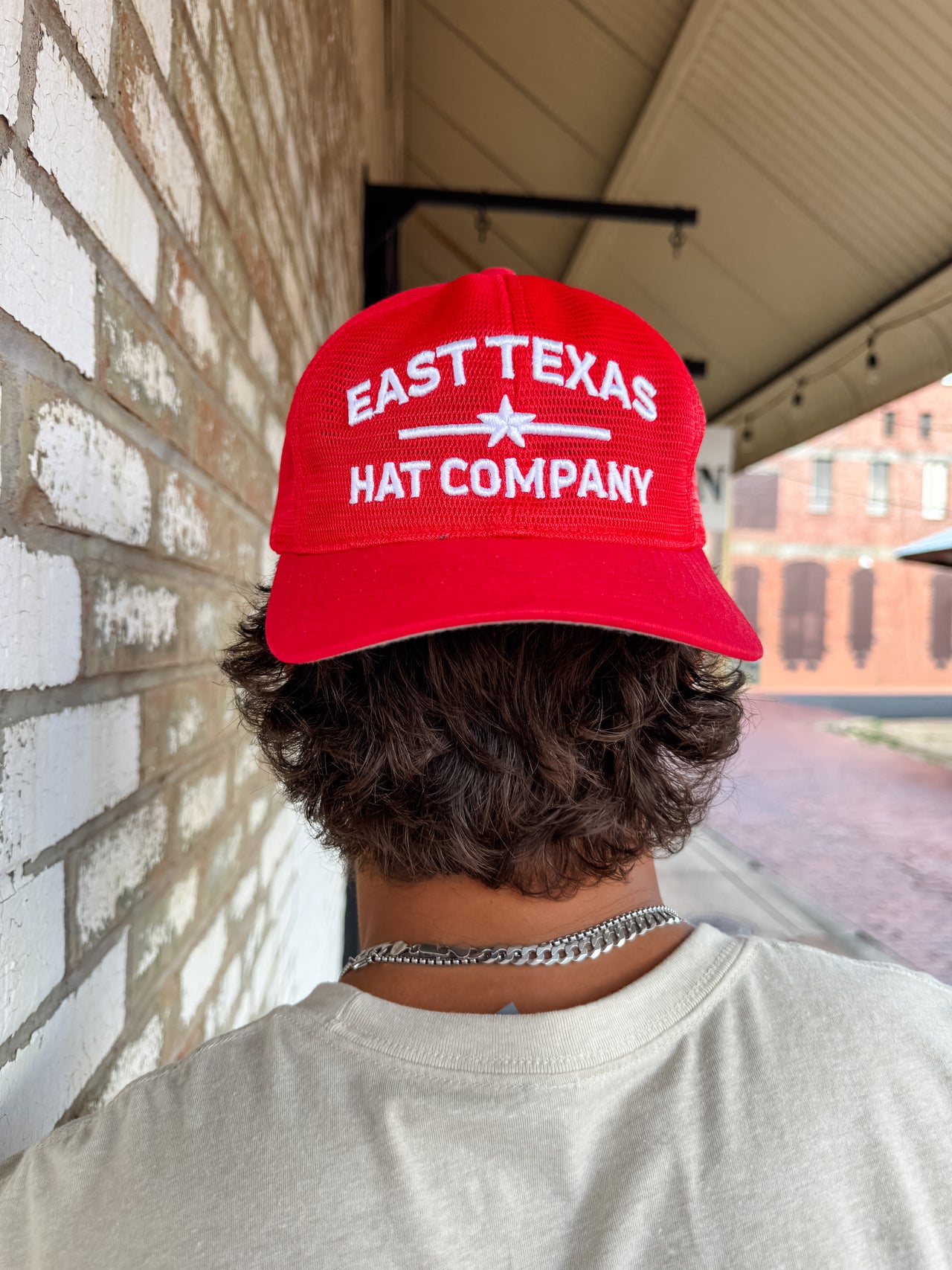 A close-up from behind of a person with wavy brown hair wearing a bright red "East Texas Hat Company" mesh trucker hat backwards. The hat features white embroidered block text separated by a star graphic.