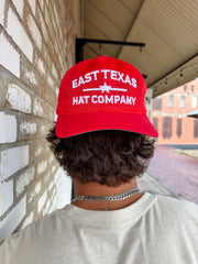 A close-up from behind of a person with wavy brown hair wearing a bright red "East Texas Hat Company" mesh trucker hat backwards. The hat features white embroidered block text separated by a star graphic.