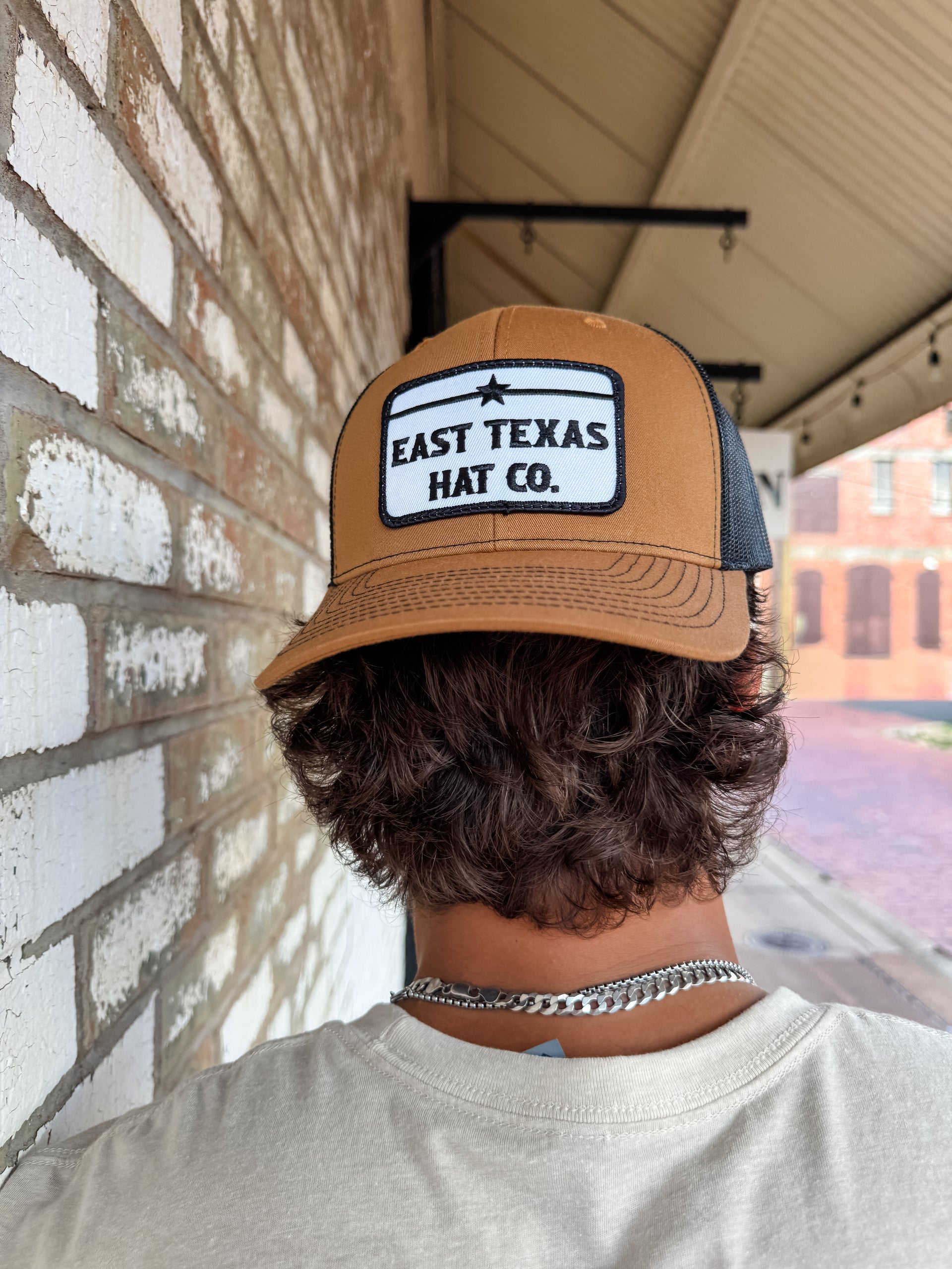 Person wearing a tan and black cap with 'East Texas Hat Co.' logo against a brick wall.