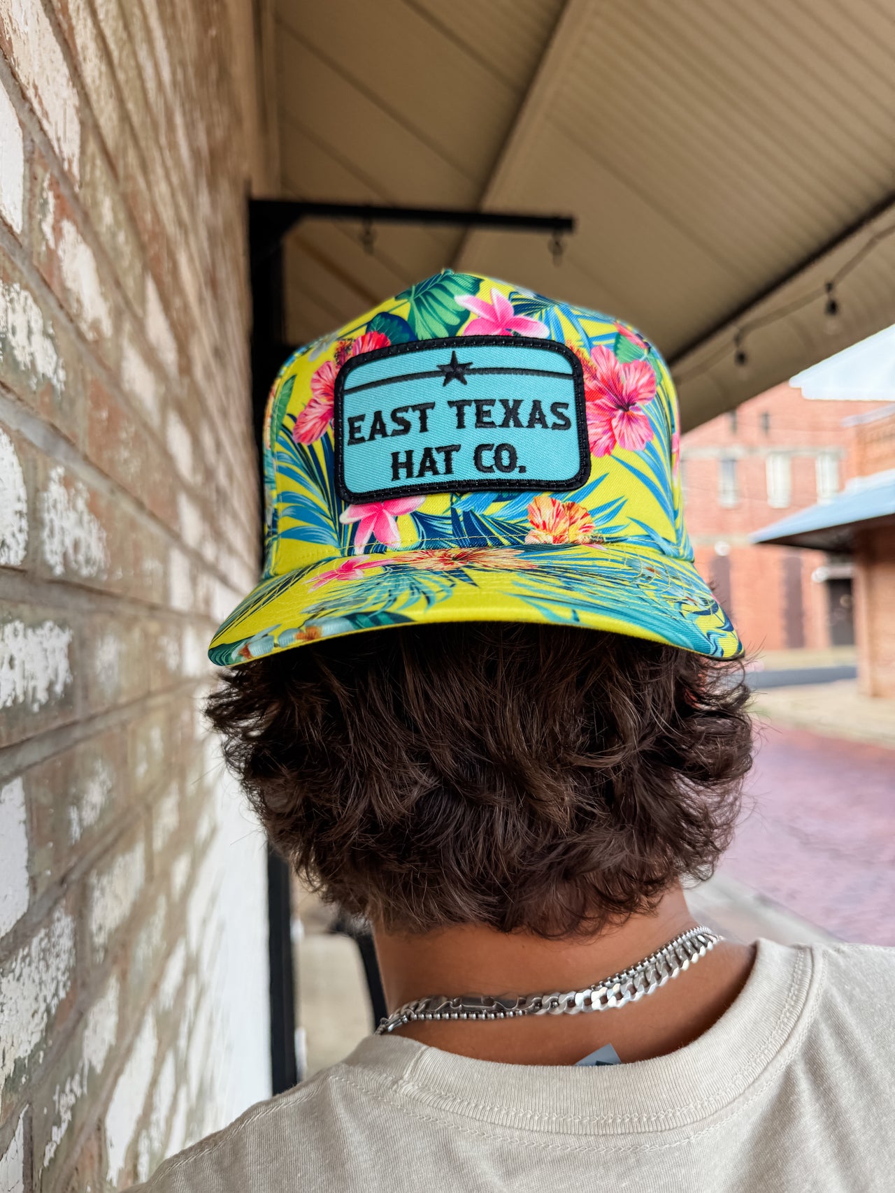 Person wearing a colorful 'East Texas Hat Co.' cap with floral design against a brick wall.