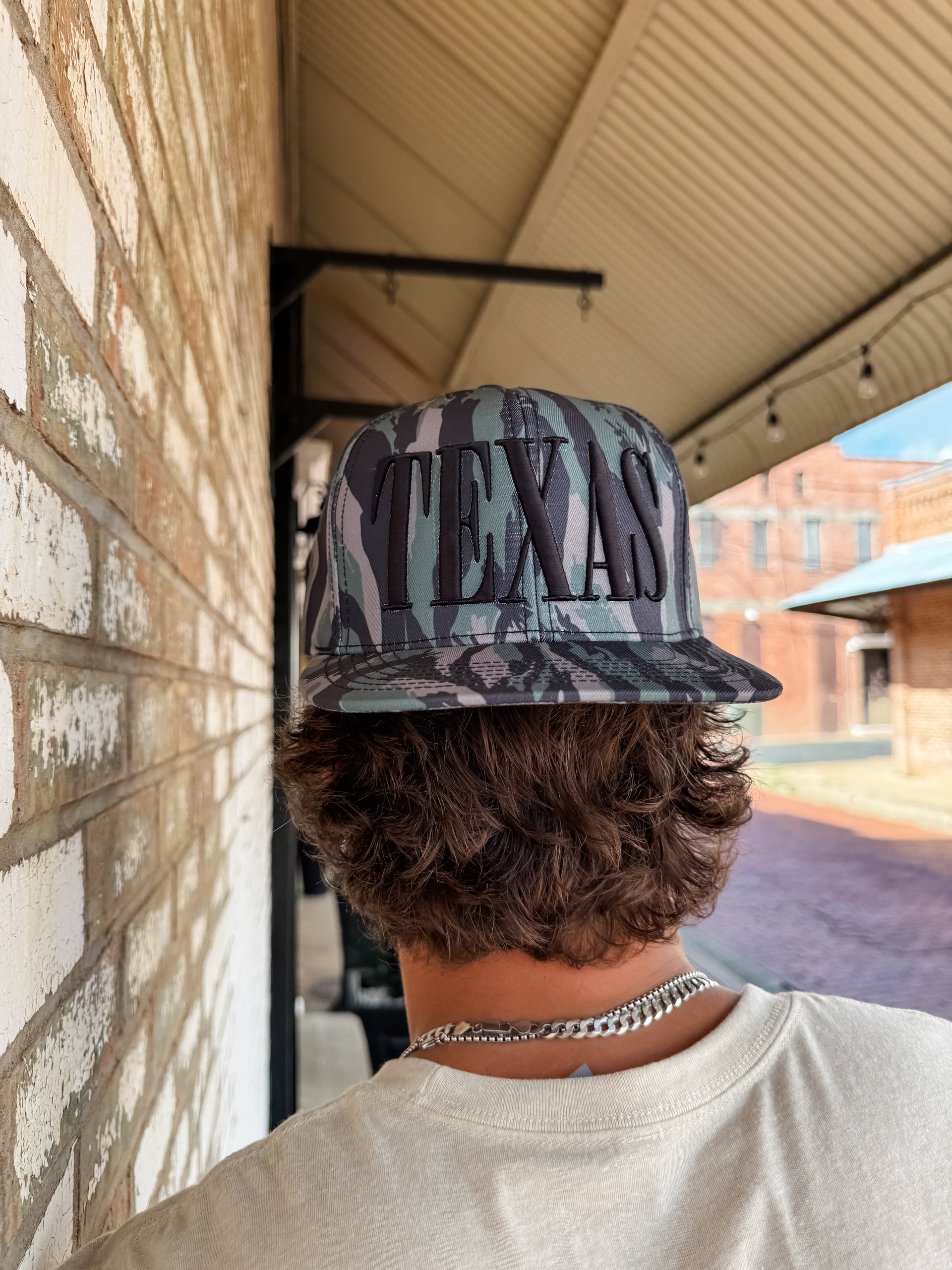 Person wearing a 'Texas' cap with camouflage design, standing against a brick wall.