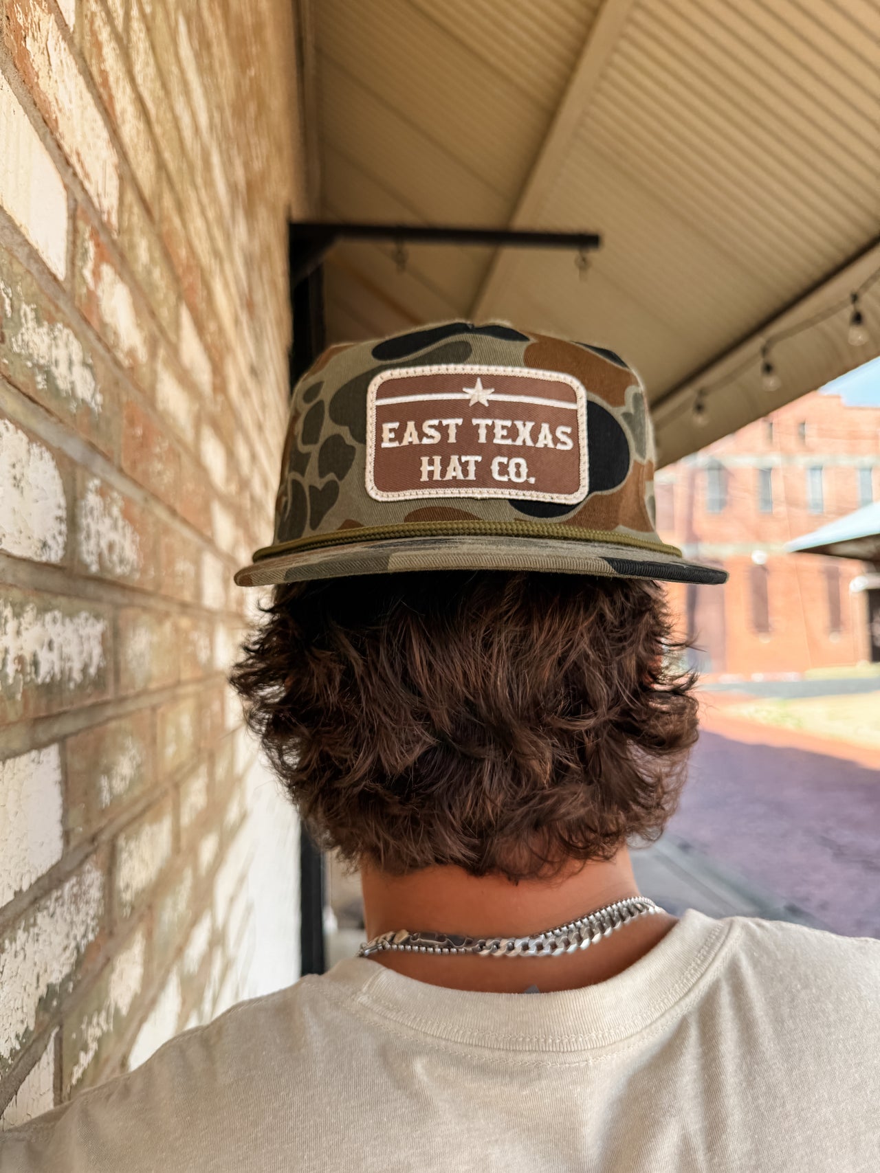 Person wearing a 'East Texas Hat Co.' cap with a brick wall and building in the background