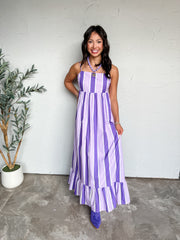 A full-length photograph of a smiling woman with long dark wavy hair posing with one hand on her hip against a textured white wall next to a potted olive tree. She is wearing a sleeveless maxi dress with thin spaghetti straps featuring vertical stripes in deep purple, lavender, and white made from 100% cotton, with a square neck, an elastic Empire waist, and a ruffled tier at the hem, paired with purple suede cowboy boots, styled with a light purple beaded necklace featuring a gold star pendant and delicate