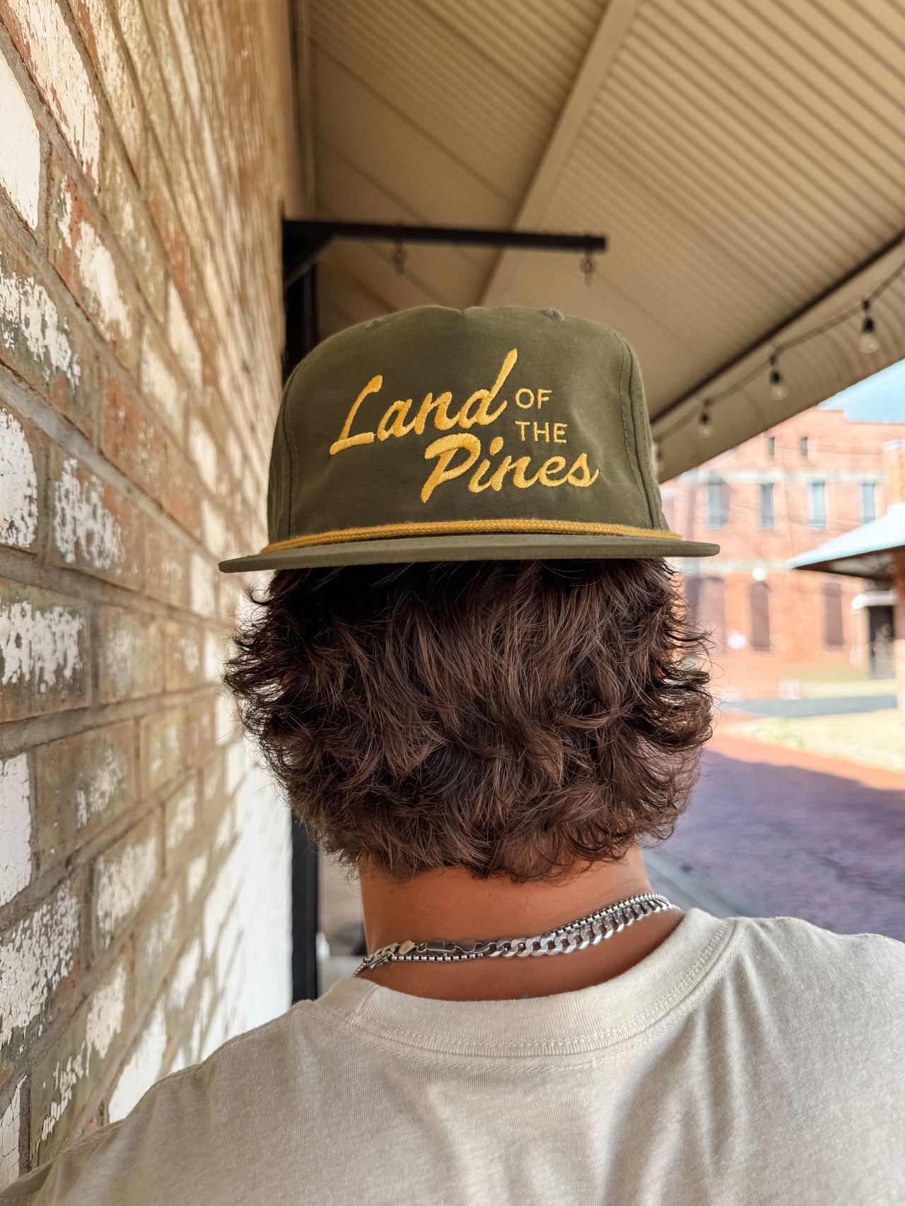Person wearing a cap with 'Land of the Pines' text, standing against a brick wall.