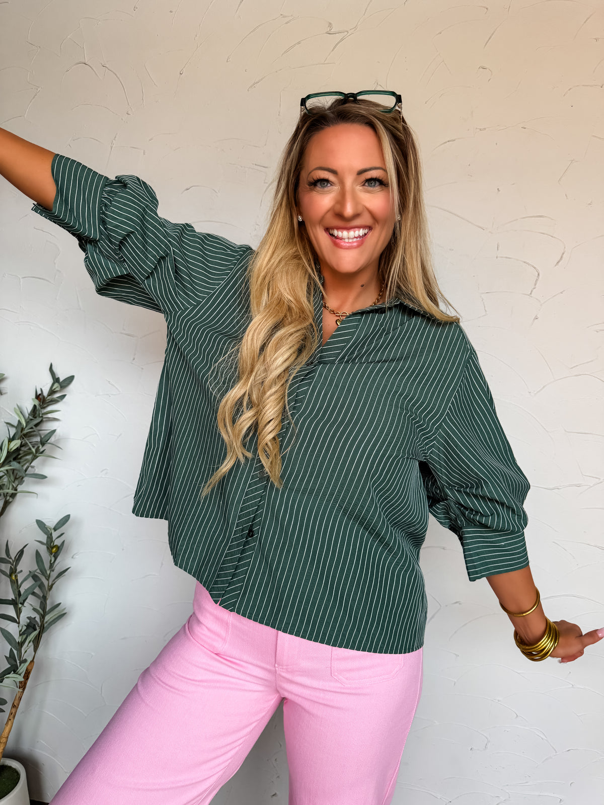 A waist-up photograph of a smiling blonde woman with glasses resting on top of her head, posing with her arms outstretched against a textured white wall. She is wearing a three-quarter sleeve button-down shirt made from a 65% polyester and 35% cotton blend with forest green and white vertical pinstripes, featuring a pointed collar and relaxed sleeves with cuffs, paired with light pink pants and styled with stacked gold bangles and a gold chain necklace.