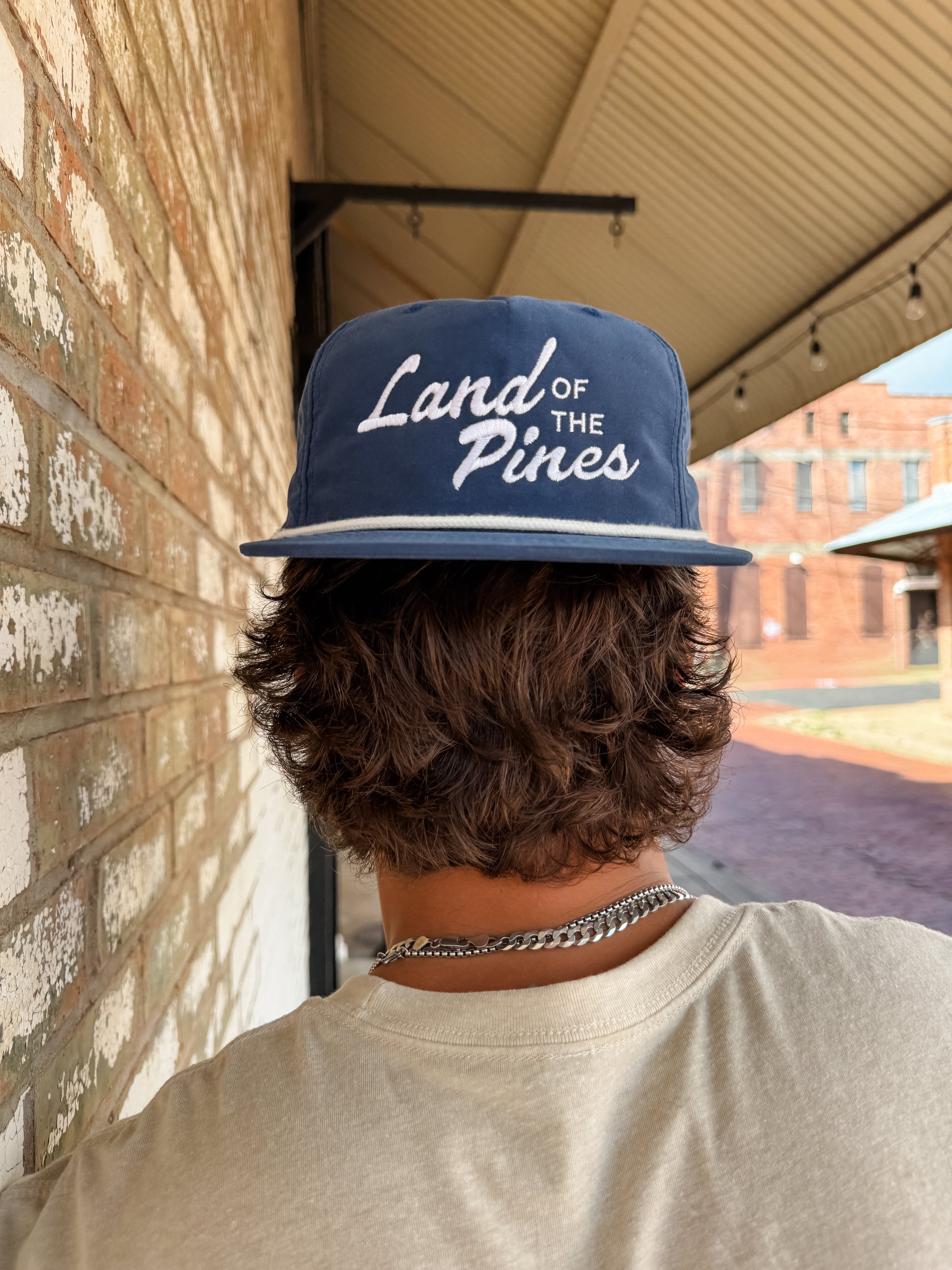 Person wearing a blue cap with 'Land of the Pines' text, standing in front of a brick wall.