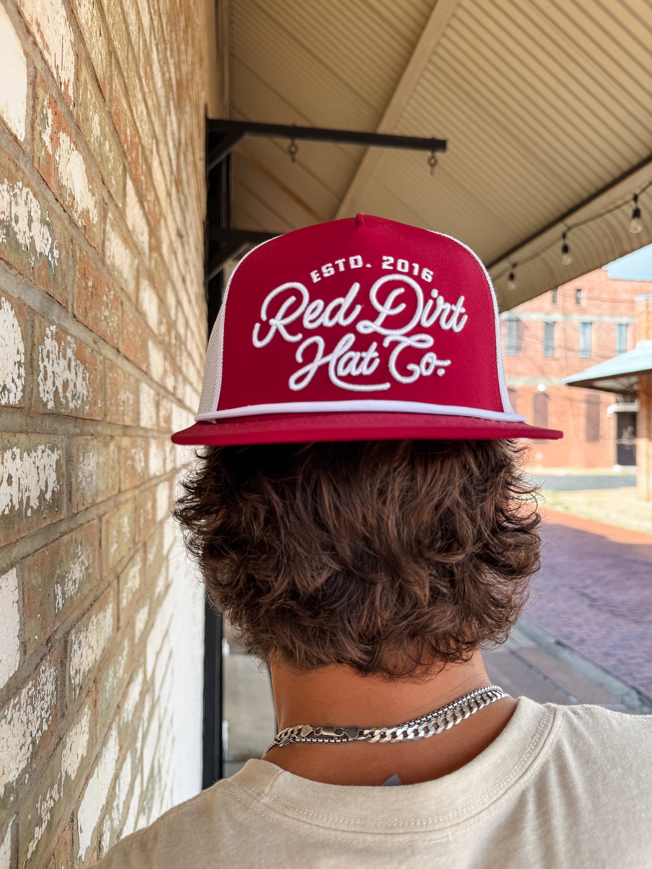 Person wearing a red 'Red Dirt Hat Co' cap in front of a brick wall.