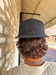 A close-up from behind of a person with wavy brown hair wearing an all-black "Red Dirt Hat Co." trucker hat backwards. The hat features a black-on-black embossed "RDH Co." monogram logo on the front and a black mesh back.
