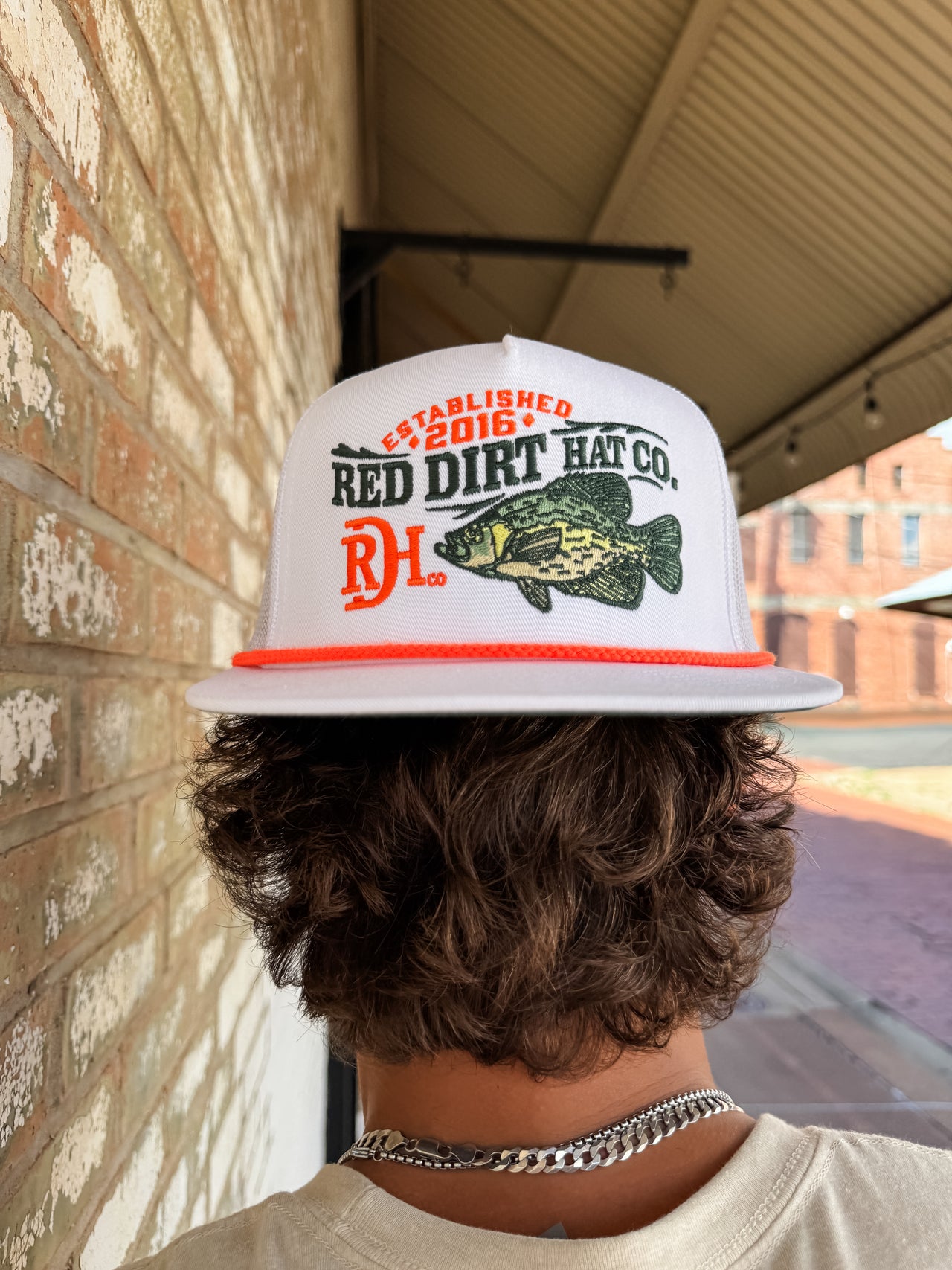 Person wearing a 'Red Dirt Hat Co.' cap with a fish graphic, standing against a brick wall.