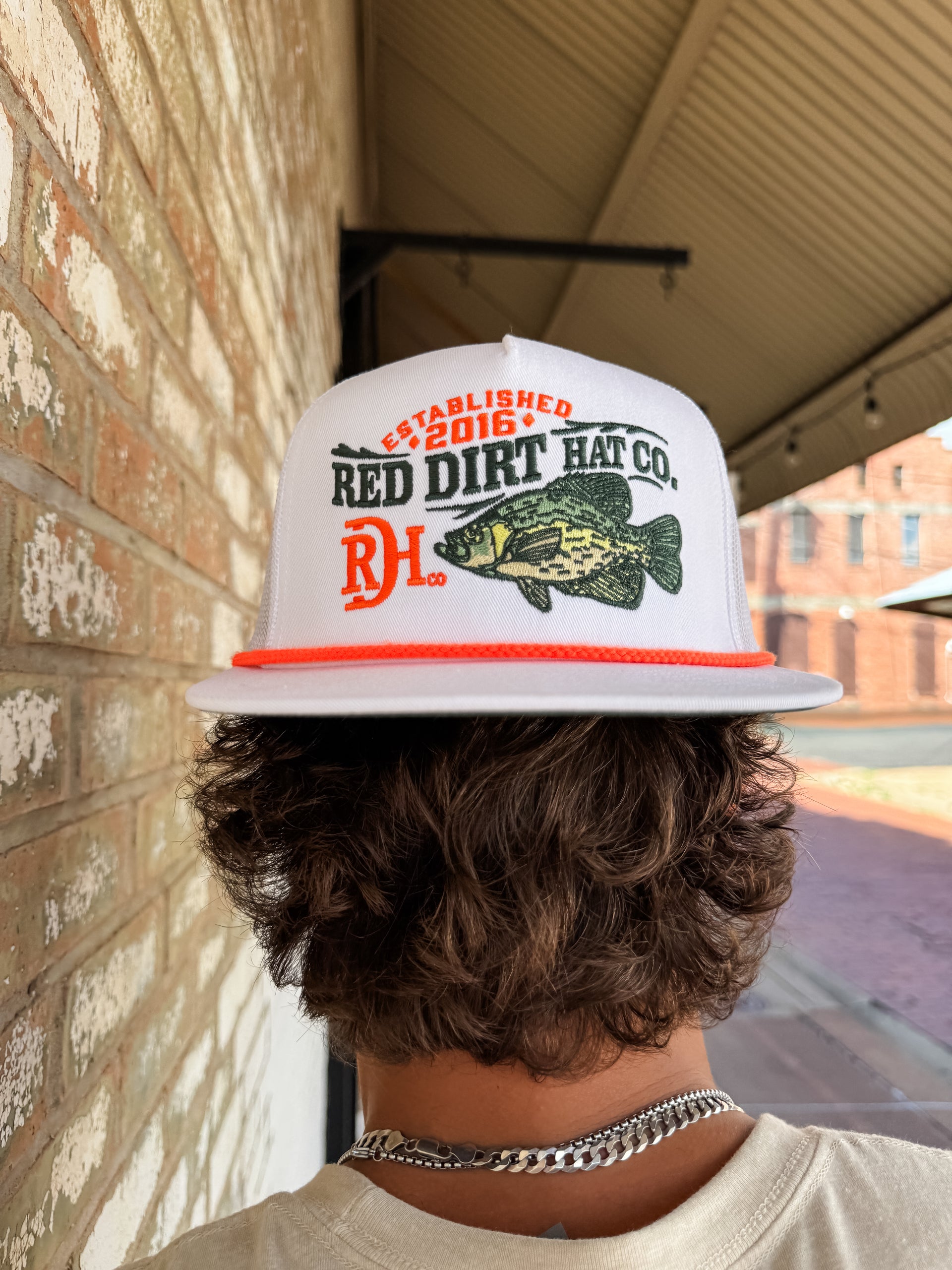 Person wearing a 'Red Dirt Hat Co.' cap with a fish graphic, standing against a brick wall.