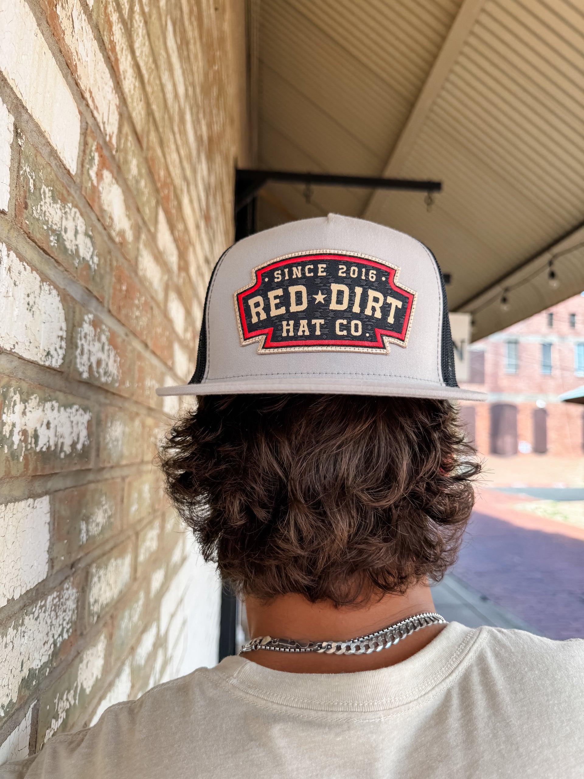 A close-up from behind of a person with wavy brown hair wearing a light gray "Red Dirt Hat Co." trucker hat backwards. The hat features a black, red, and tan embroidered shield-shaped patch and a black mesh back.