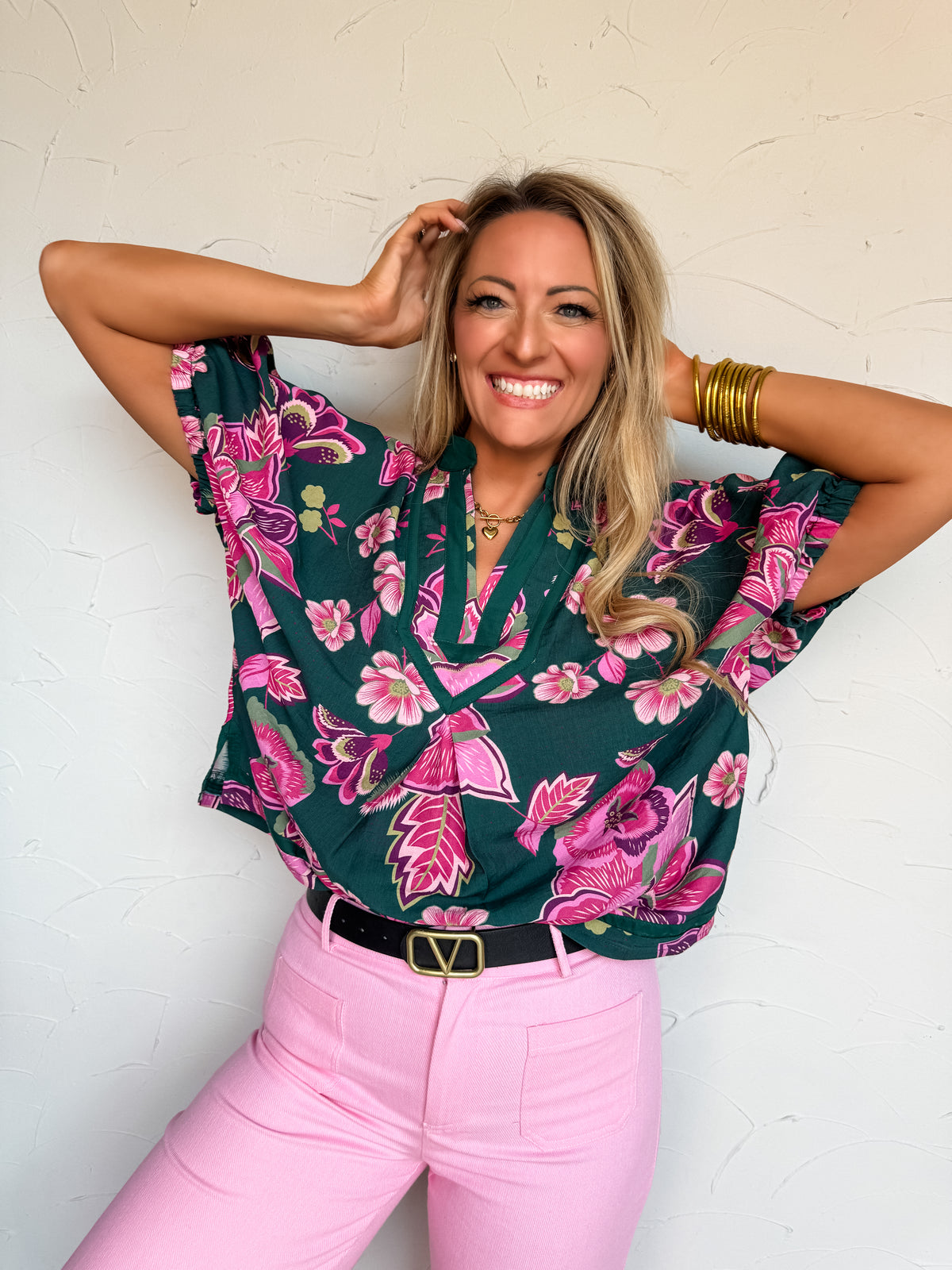 A waist-up photograph of a smiling blonde woman posing with both hands behind her head against a textured white wall. She is wearing a short-sleeved blouse made from an 88% viscose and 12% nylon blend with a bold magenta pink and purple floral pattern on a dark emerald green base, featuring a notched V-neckline with decorative stitched trim and wide short sleeves, tucked into light pink pants with a black belt, and styled with stacked gold bangles and a gold chain heart necklace.