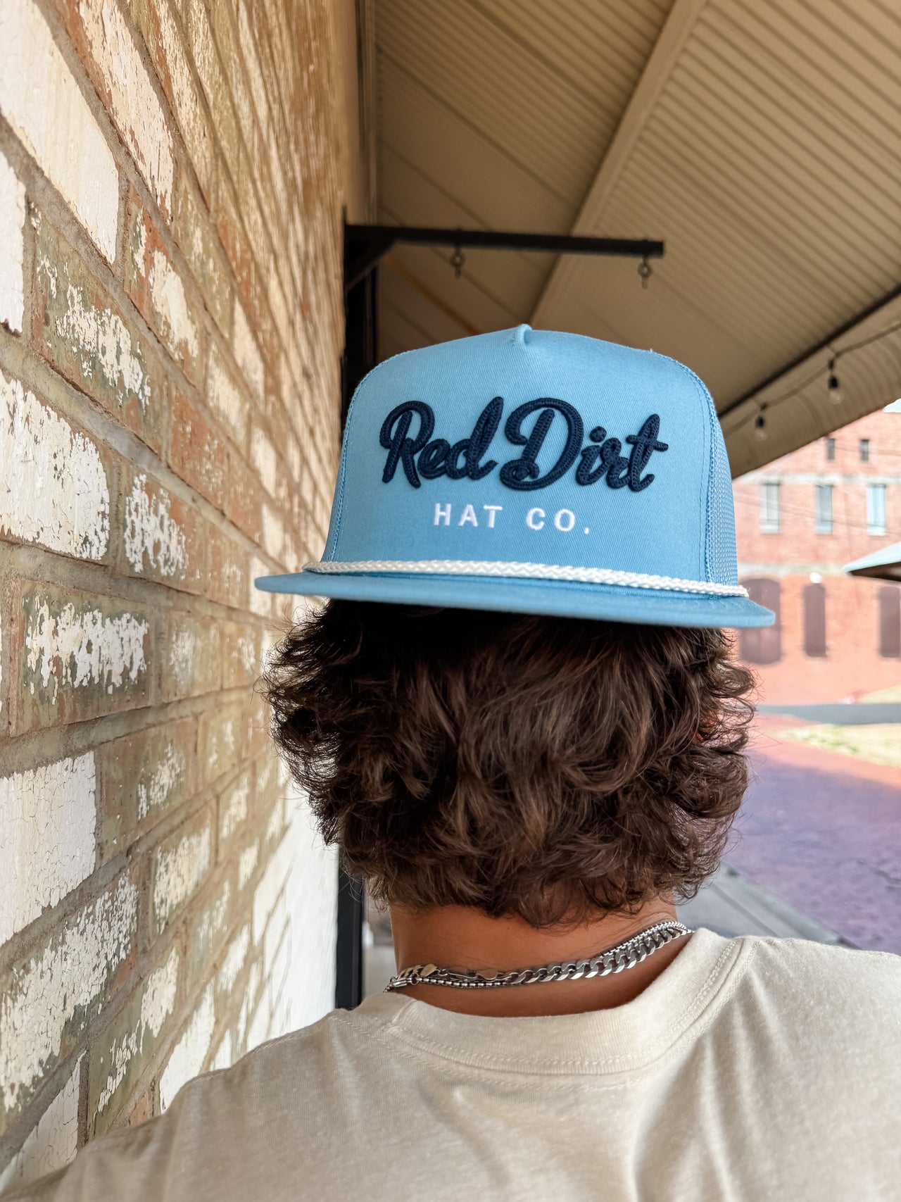A close-up from behind of a person with wavy brown hair wearing a light blue "Red Dirt Hat Co." trucker hat backwards. The hat features dark blue embroidered cursive text, white block lettering, and a white rope detail along the brim.