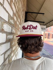 A close-up from behind of a person with wavy brown hair wearing a cream-colored "Red Dirt Hat Co." trucker hat backwards. The hat features maroon embroidered cursive text, a maroon brim, and a gold rope detail.