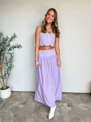 A full-length photograph of a smiling young woman with long wavy brown hair with blonde highlights posing against a textured white wall next to a potted olive tree. She is wearing a two-piece lavender and white polka dot set made from 100% polyester, featuring a crop top with thin shoulder straps, scalloped edges, and white geometric embroidery, paired with a matching high-waisted maxi skirt with an embroidered waistband, styled with white pointed-toe ankle boots, a pearl necklace with a gold heart pendant,