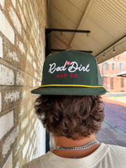 A close-up from behind of a person with wavy brown hair wearing a dark green "Red Dirt Hat Co." trucker hat backwards. The hat features white cursive text, a small red golf flag icon, and a yellow rope detail along the brim.