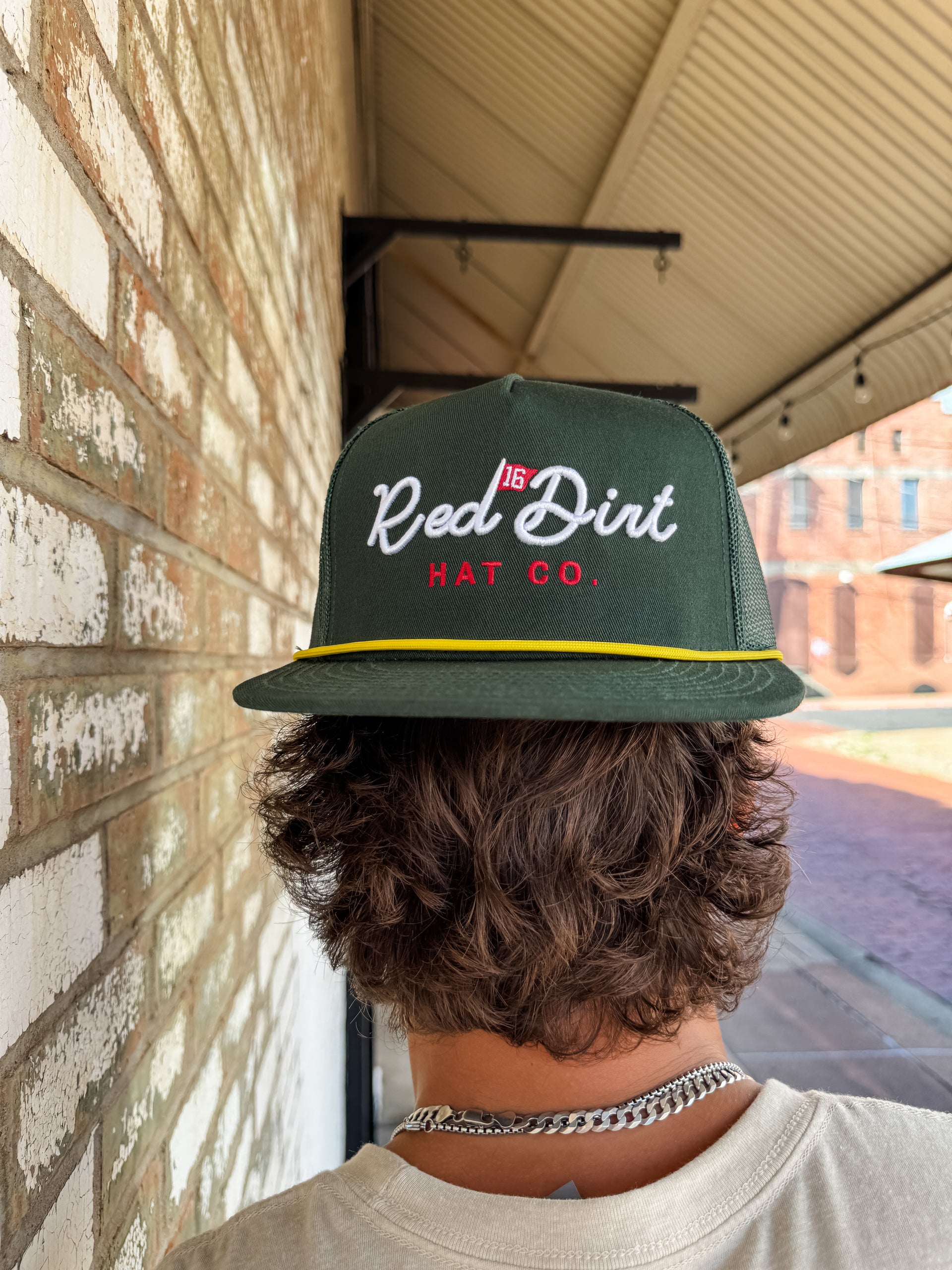 A close-up from behind of a person with wavy brown hair wearing a dark green "Red Dirt Hat Co." trucker hat backwards. The hat features white cursive text, a small red golf flag icon, and a yellow rope detail along the brim.