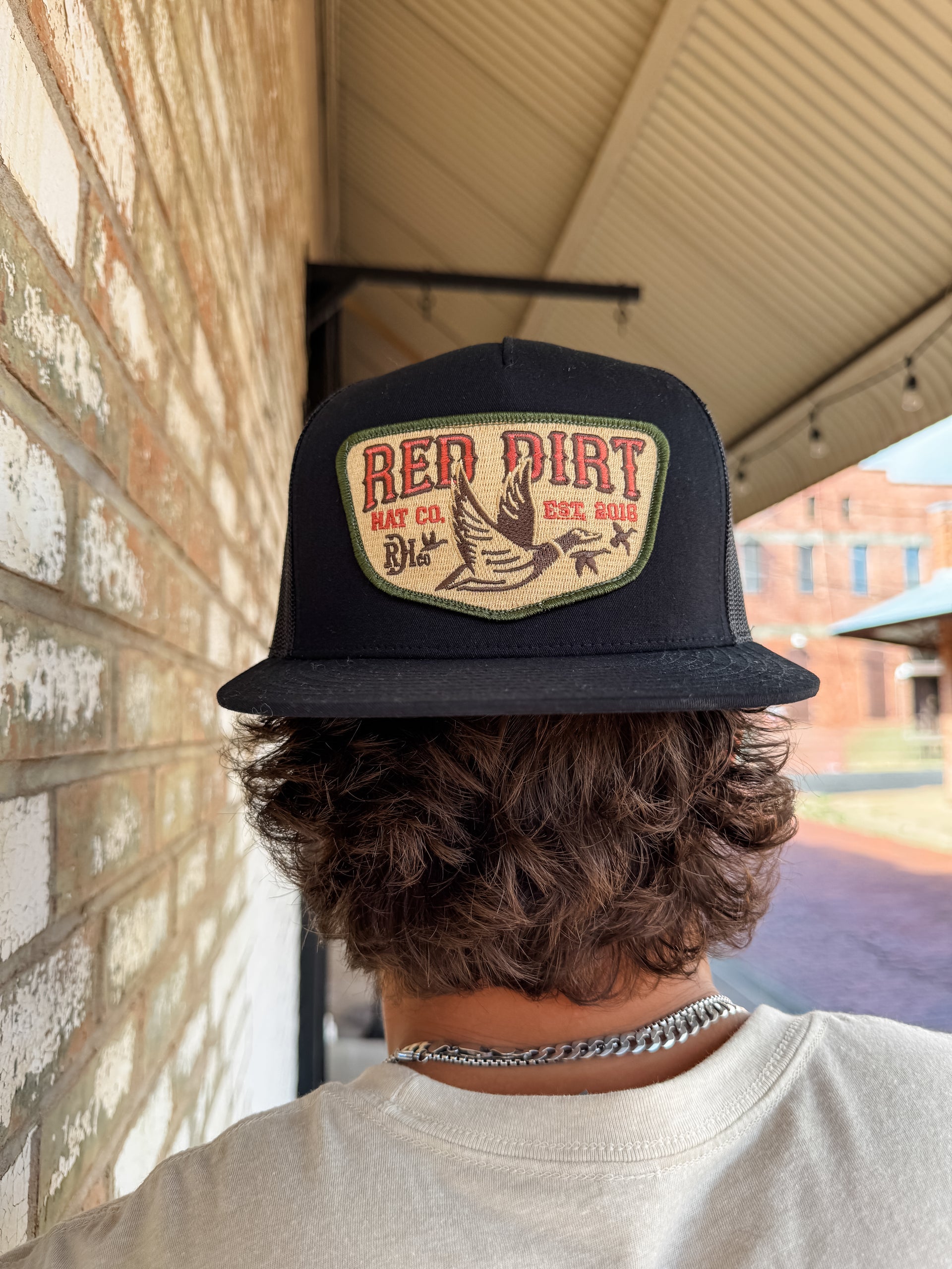 Person wearing a black cap with 'Red Dirt Hat Co' logo, standing in front of a brick wall.