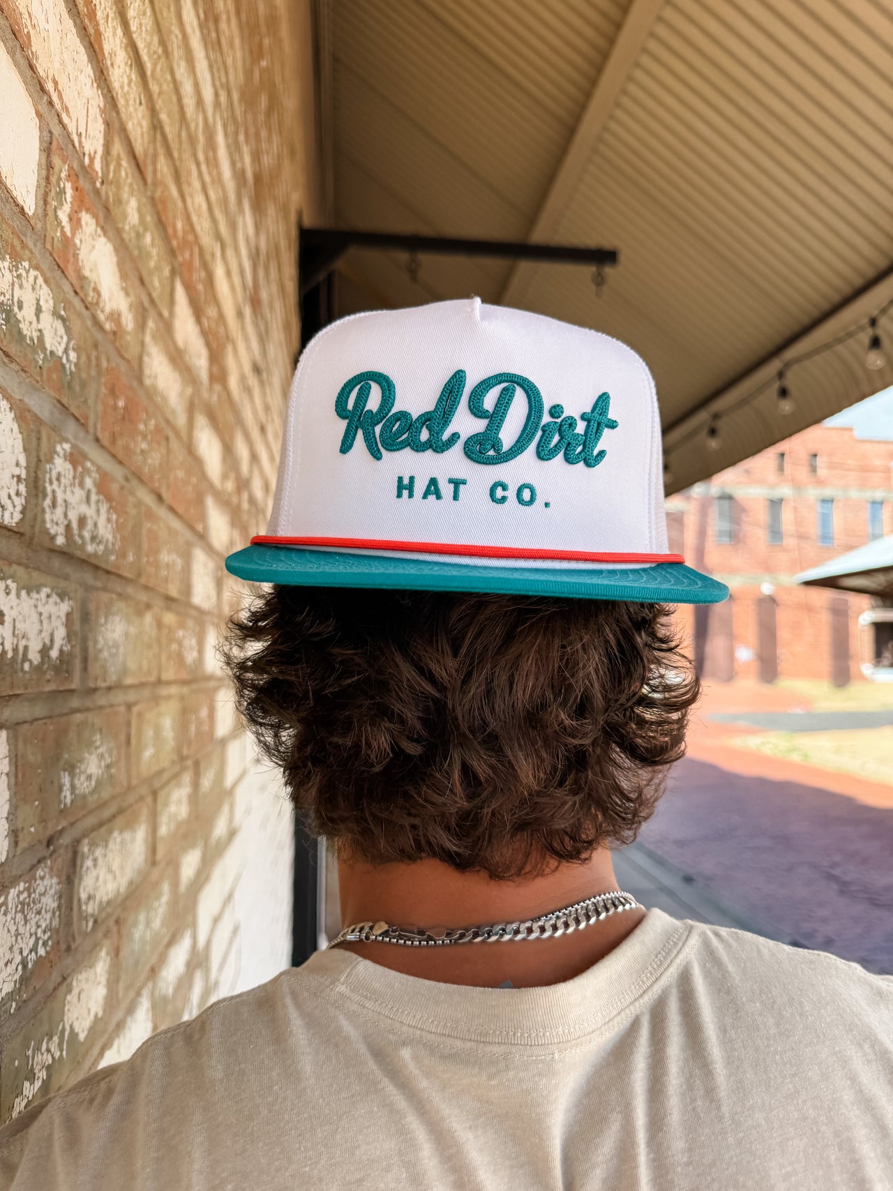 Person wearing a 'Red Dirt Hat Co.' cap with a brick wall and outdoor setting in the background