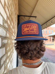 Person wearing a navy blue cap with orange text and logo, standing against a brick wall.