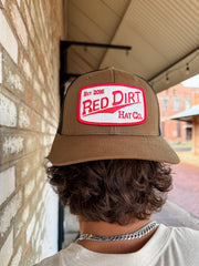 Person wearing a brown cap with a 'Red Dirt Hat Co' logo, standing against a brick wall.