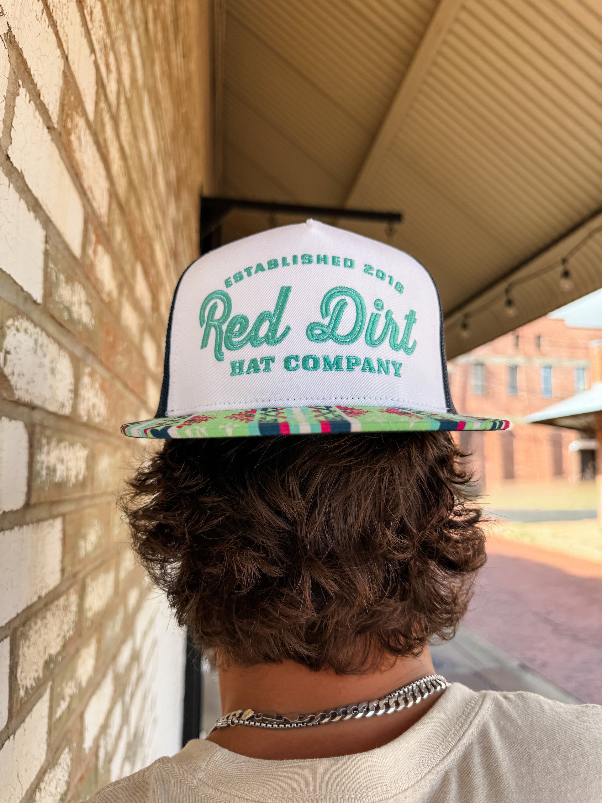 Person wearing a 'Red Dirt Hat Company' cap with a colorful brim, standing against a brick wall.
