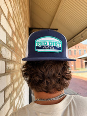 A close-up from behind of a person with wavy brown hair wearing a navy blue "Red Dirt Hat Co." trucker hat backwards. The hat features a white, teal, and navy embroidered logo patch and a white mesh back.