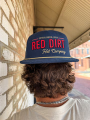 Person wearing a navy blue cap with 'Red Dirt Hat Company' text, standing against a brick wall.