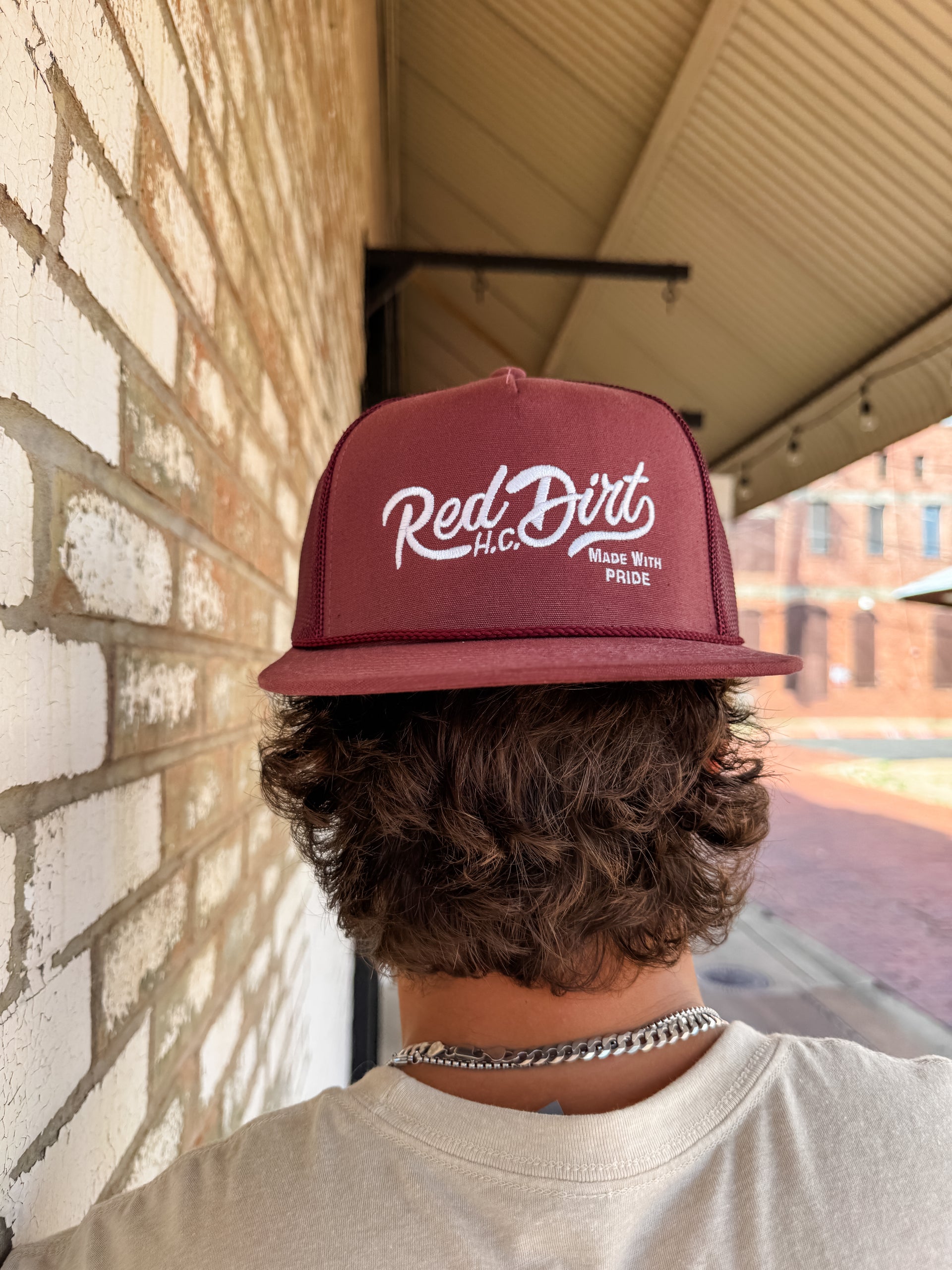 A close-up from behind of a person with wavy brown hair wearing a maroon "Red Dirt H.C." trucker hat backwards. The hat features white embroidered cursive text, the words "Made With Pride," and a matching maroon mesh back.