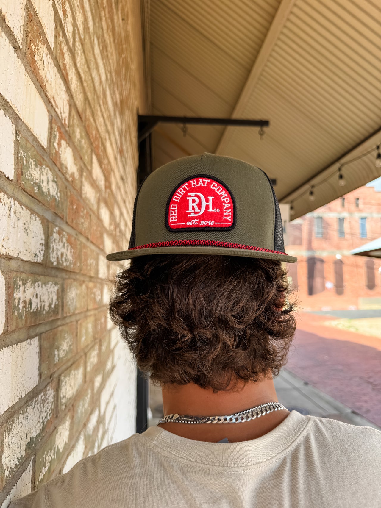 A close-up from behind of a person with wavy brown hair wearing an olive green "Red Dirt Hat Company" trucker hat backwards. The hat features a red and white circular embroidered logo patch, a patterned red and black rope detail along the brim, and a black mesh back.