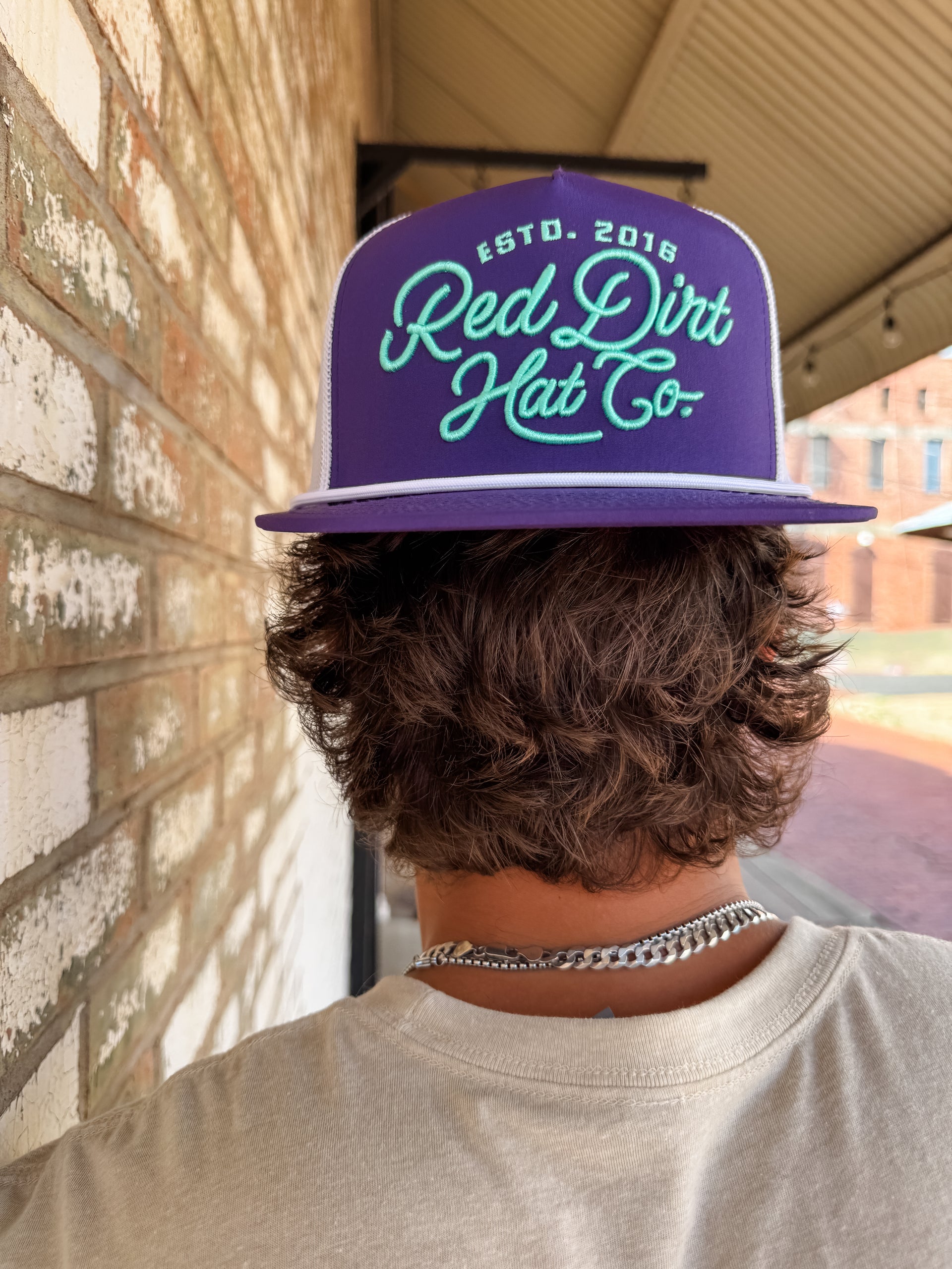 A close-up from behind of a person with wavy brown hair wearing a purple "Red Dirt Hat Co." trucker hat backwards. The hat features mint green embroidered script text and a white mesh back.