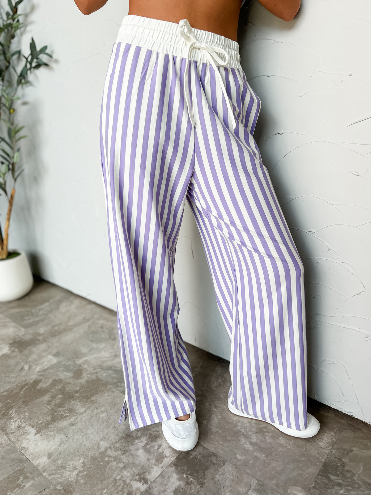 A person wearing loose, wide-leg striped pants with vertical purple and white stripes, a white elastic drawstring waist, and white slip-on shoes, standing against a light wall.